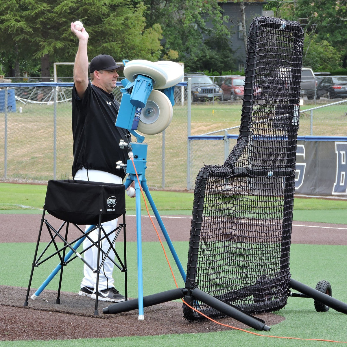A coach feeding a JUGS three-wheel pitching machine behind the JUGS Protector Series L-Shaped Screen on a baseball field.