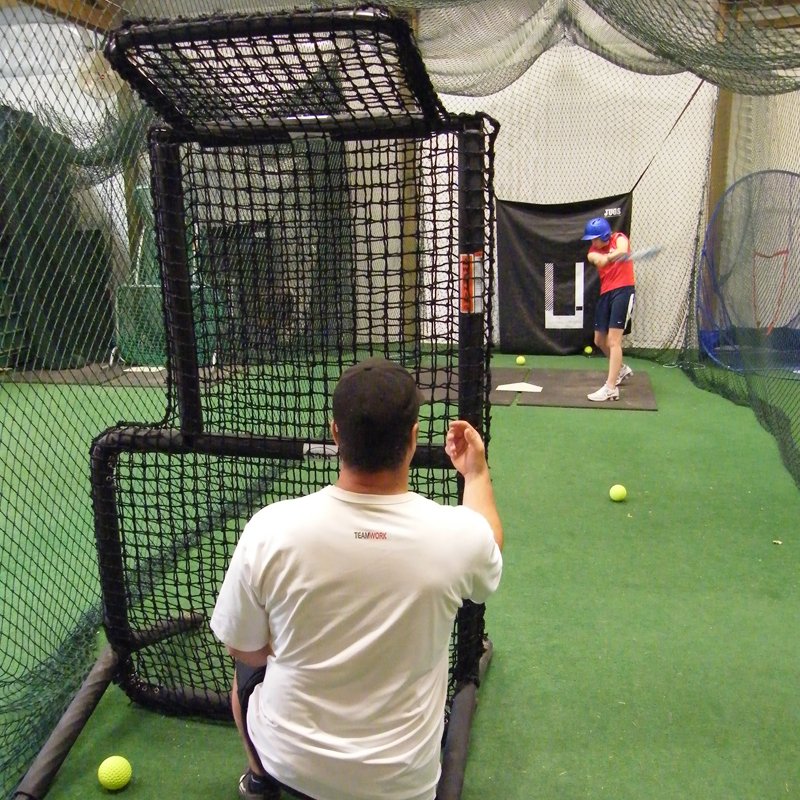 A coach tossing to a hitter from behind the JUGS Protector Series Short-Toss Screen inside a batting cage.