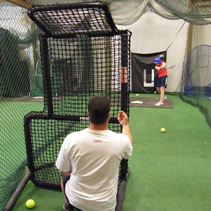 A coach tossing to a hitter from behind the JUGS Protector Series Short-Toss Screen inside a batting cage.