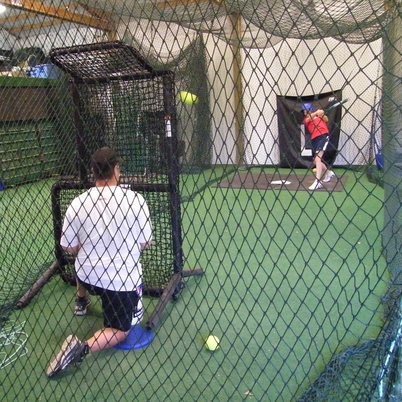 A coach kneeling behind the JUGS Protector Series Short-Toss Screen as a hitter swings in an indoor cage.