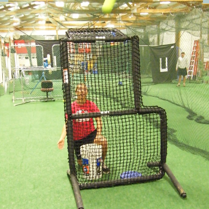 A coach seated behind the JUGS Protector Series Short-Toss Screen in an indoor batting cage.