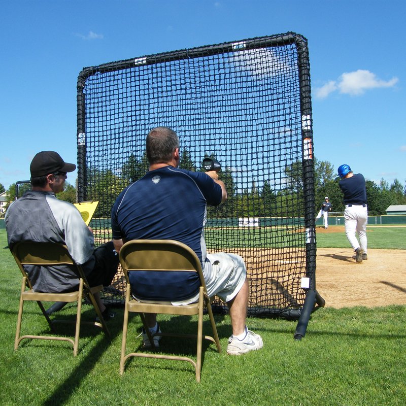 Two people seated behind the JUGS Protector Series Square Baseman Screen near a baseball diamond.
