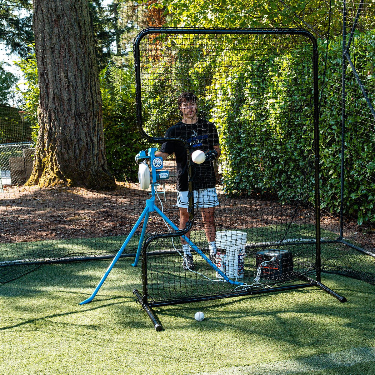 A player feeding a JUGS PS50 pitching machine on backyard turf behind a protective L-screen in a backyard batting cage.