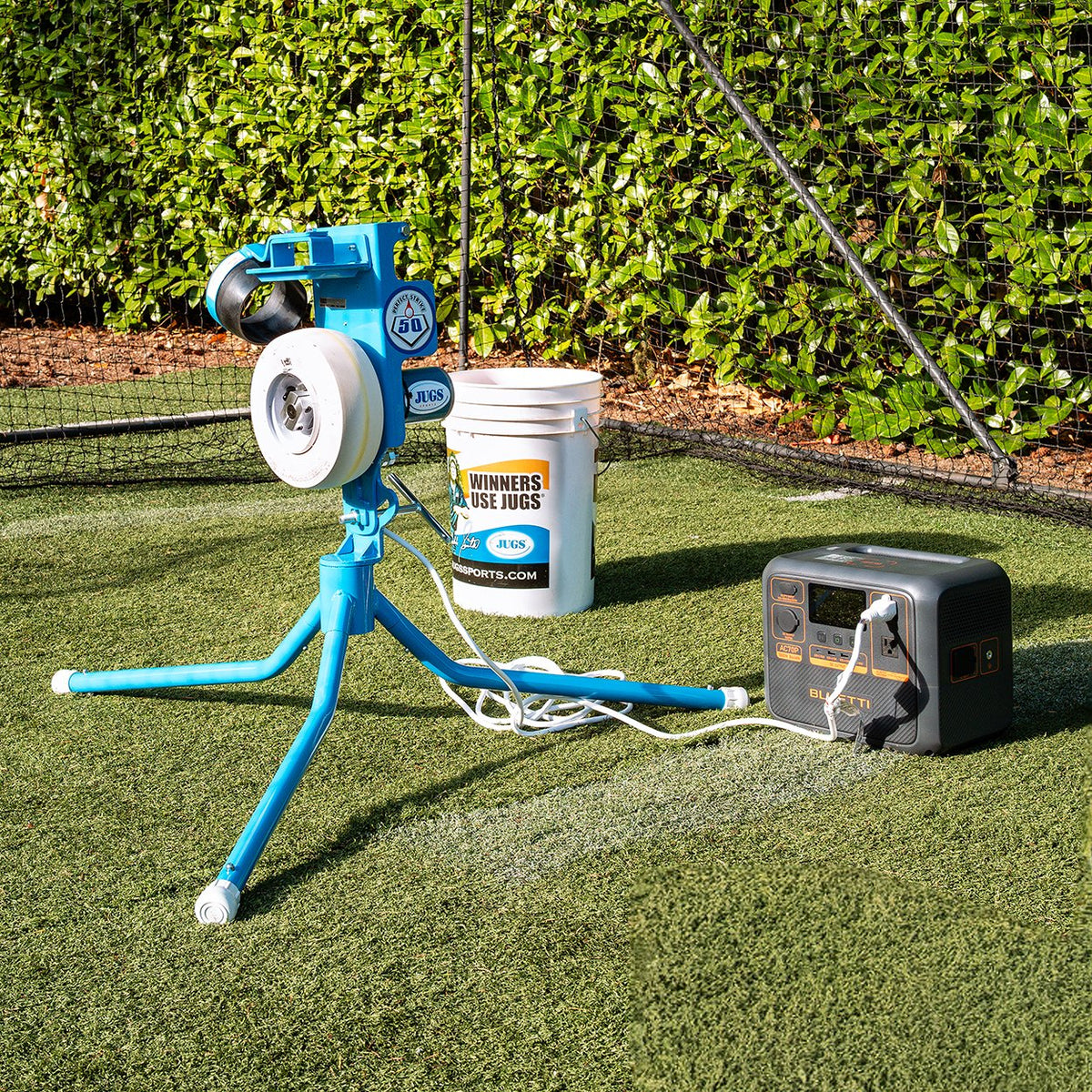 A JUGS PS50 pitching machine set up on turf next to a bucket of balls and a portable power station.