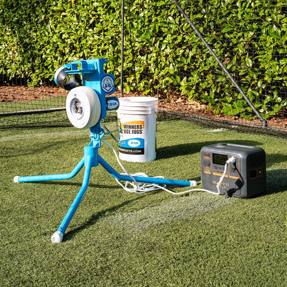 A JUGS PS50 pitching machine set up on turf next to a bucket of balls and a portable power station.