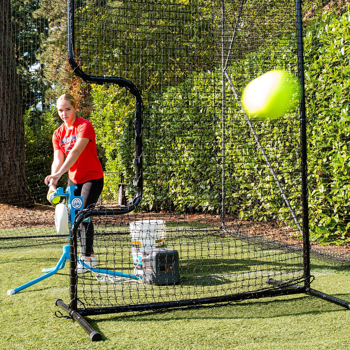 A softball player feeding a JUGS PS50 from behind a protective screen in a backyard batting cage as the ball flies toward the hitter.