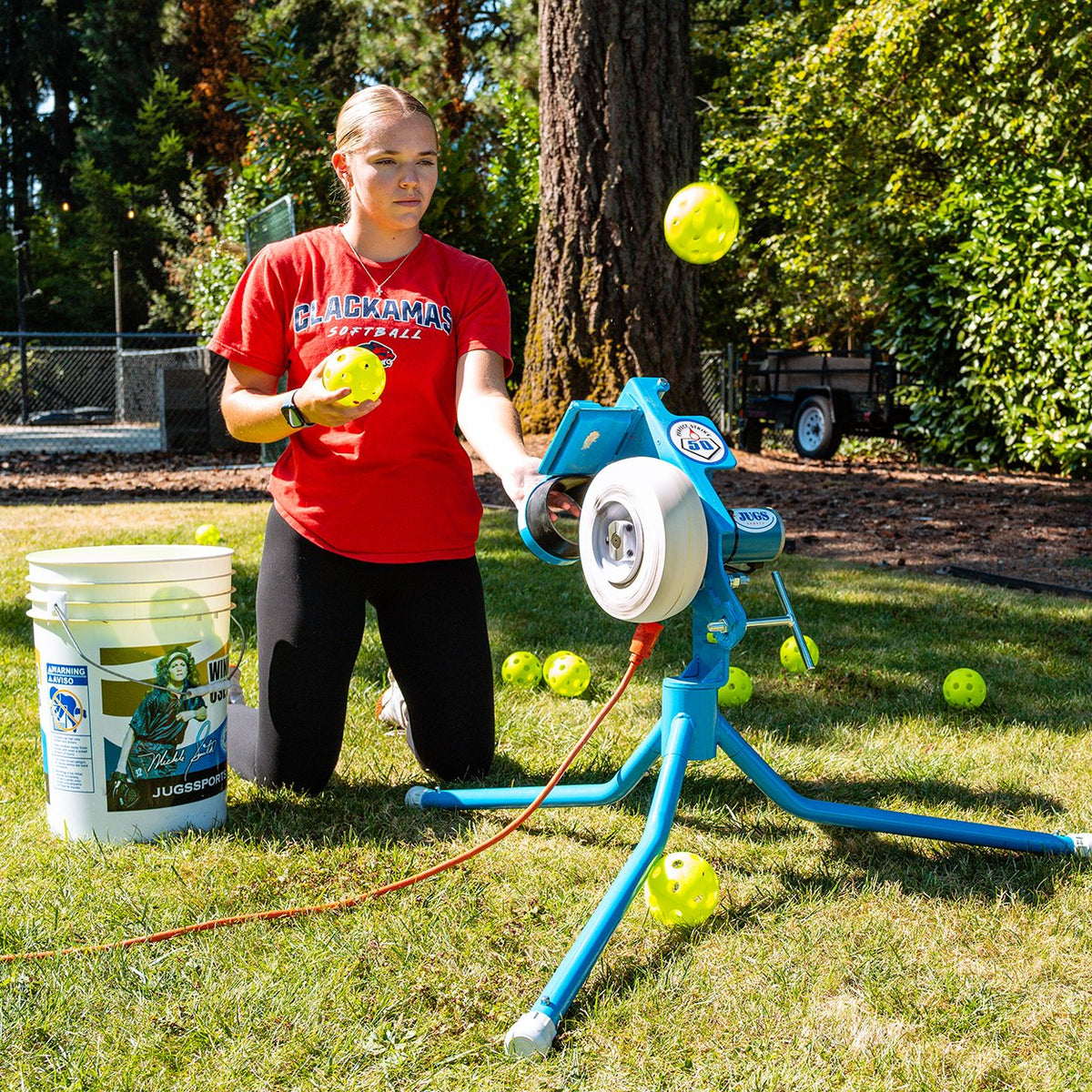 A softball player kneeling beside a JUGS PS50 on the short legs, feeding a yellow softball into the chute with a bucket of softballs nearby.