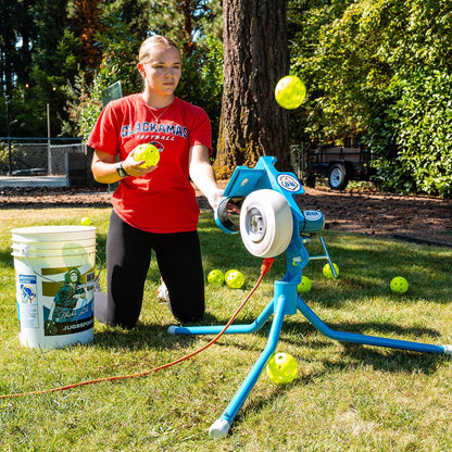 A softball player kneeling beside a JUGS PS50 on the short legs, feeding a yellow softball into the chute with a bucket of softballs nearby.