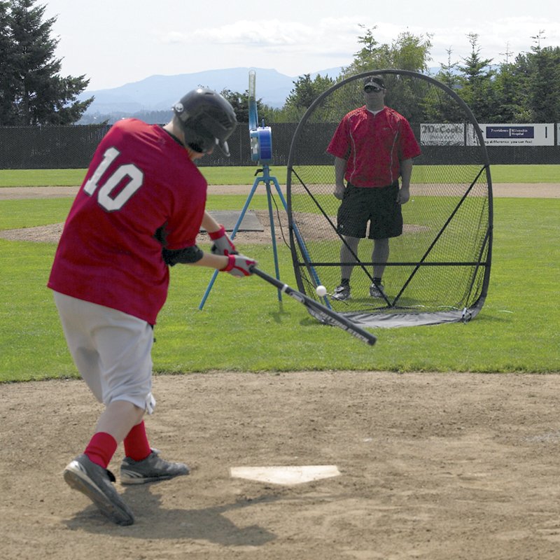 A young player in a red jersey swinging at a small ball on a dirt field, with a coach and a JUGS Small-Ball machine set in front of a net behind him.