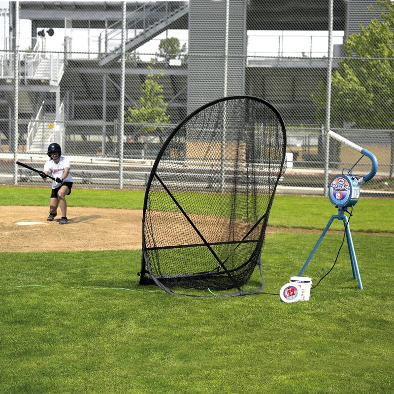A young batter at home plate on a grass field facing a JUGS Small-Ball machine set beside a black hitting net.