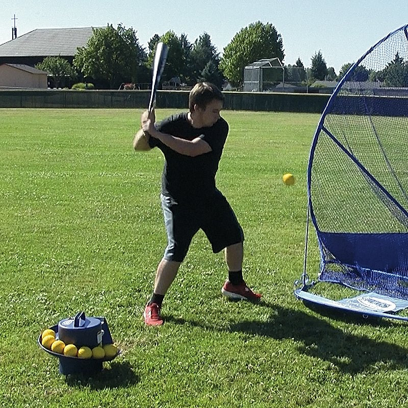 A batter in a black shirt hitting on a grass field with a JUGS Toss Machine full of yellow balls in front of him and a blue net beside him.