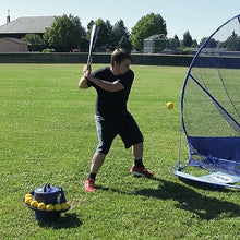 A batter in a black shirt hitting on a grass field with a JUGS Toss Machine full of yellow balls in front of him and a blue net beside him.