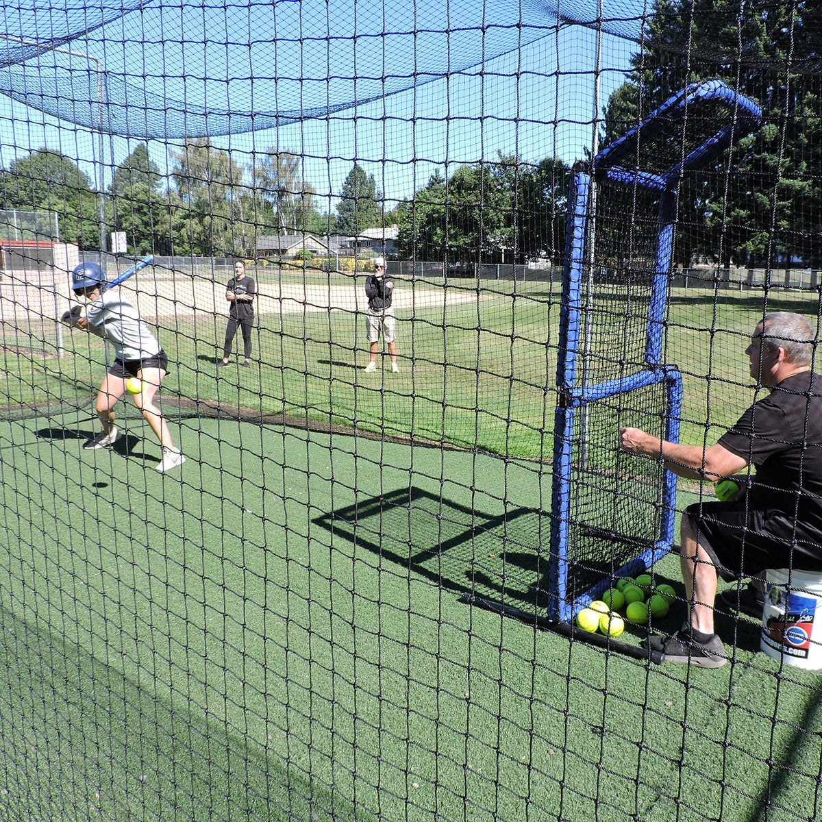 A fastpitch softball hitter in a JUGS batting cage while a coach feeds a pitching machine from behind a blue protective screen.