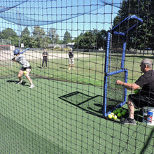 A fastpitch softball hitter in a JUGS batting cage while a coach feeds a pitching machine from behind a blue protective screen.