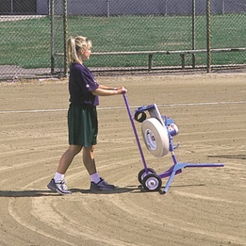 A woman wheeling a JUGS softball pitching machine across a dirt infield on the light blue transport cart.