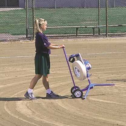 A woman wheeling a JUGS softball pitching machine across a dirt infield on the light blue transport cart.