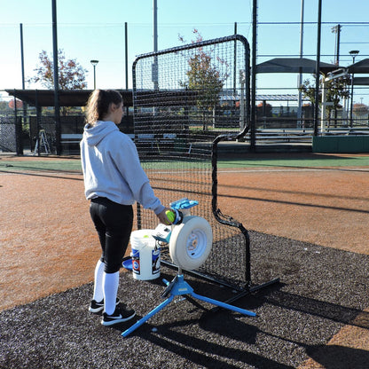 A softball player feeding a JUGS pitching machine behind the JUGS Standard C-Shaped Softball Screen.