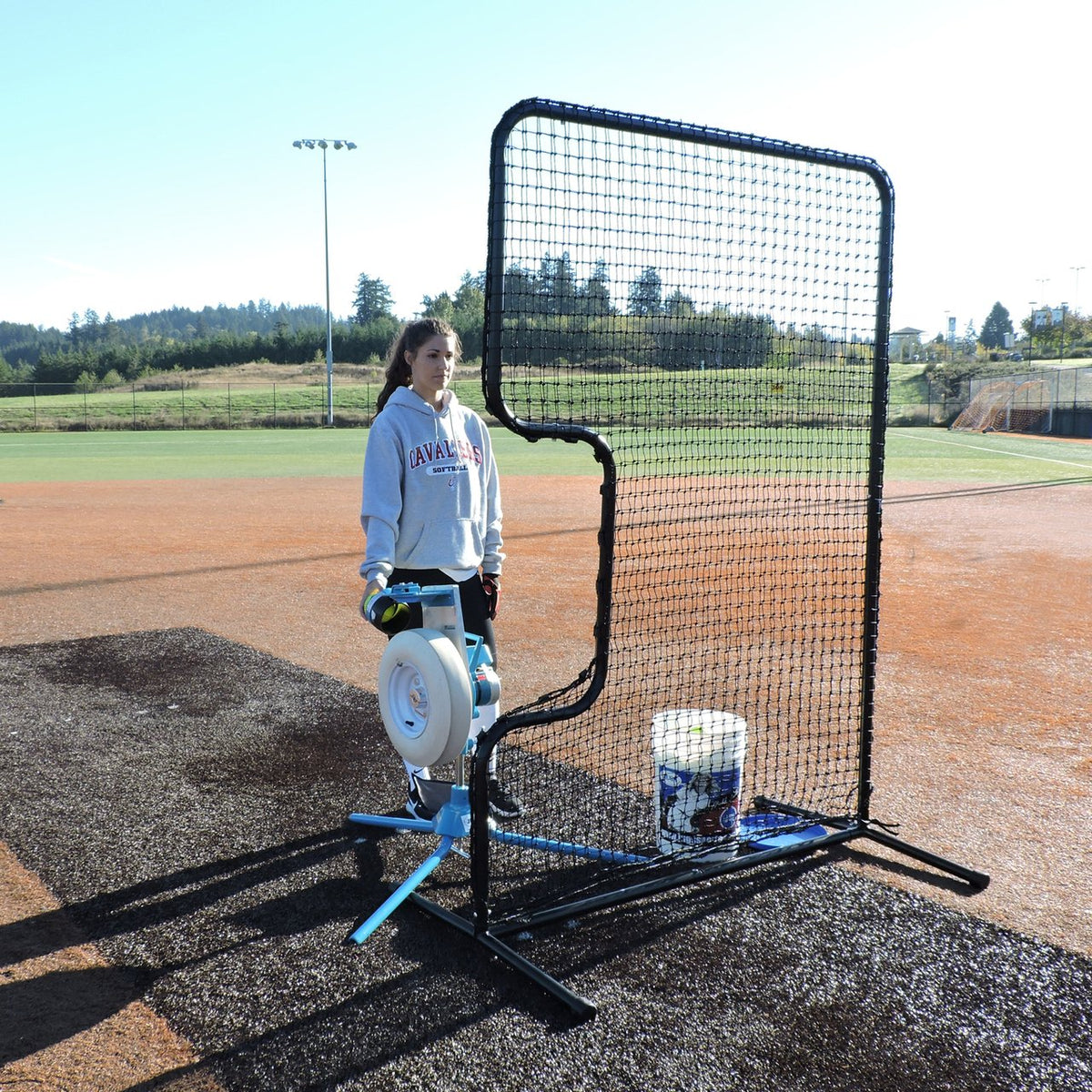 A softball player next to a JUGS pitching machine set up behind the JUGS Standard C-Shaped Softball Screen.