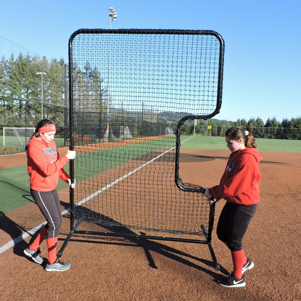 Two softball players carrying the JUGS Standard C-Shaped Softball Screen across the infield.