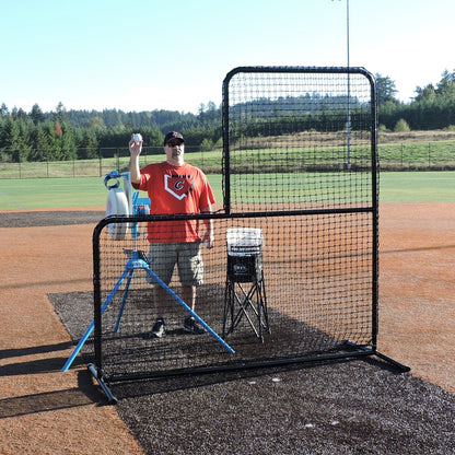 A coach feeding a JUGS pitching machine behind the JUGS Standard L-Shaped Pitchers Screen on a baseball field.