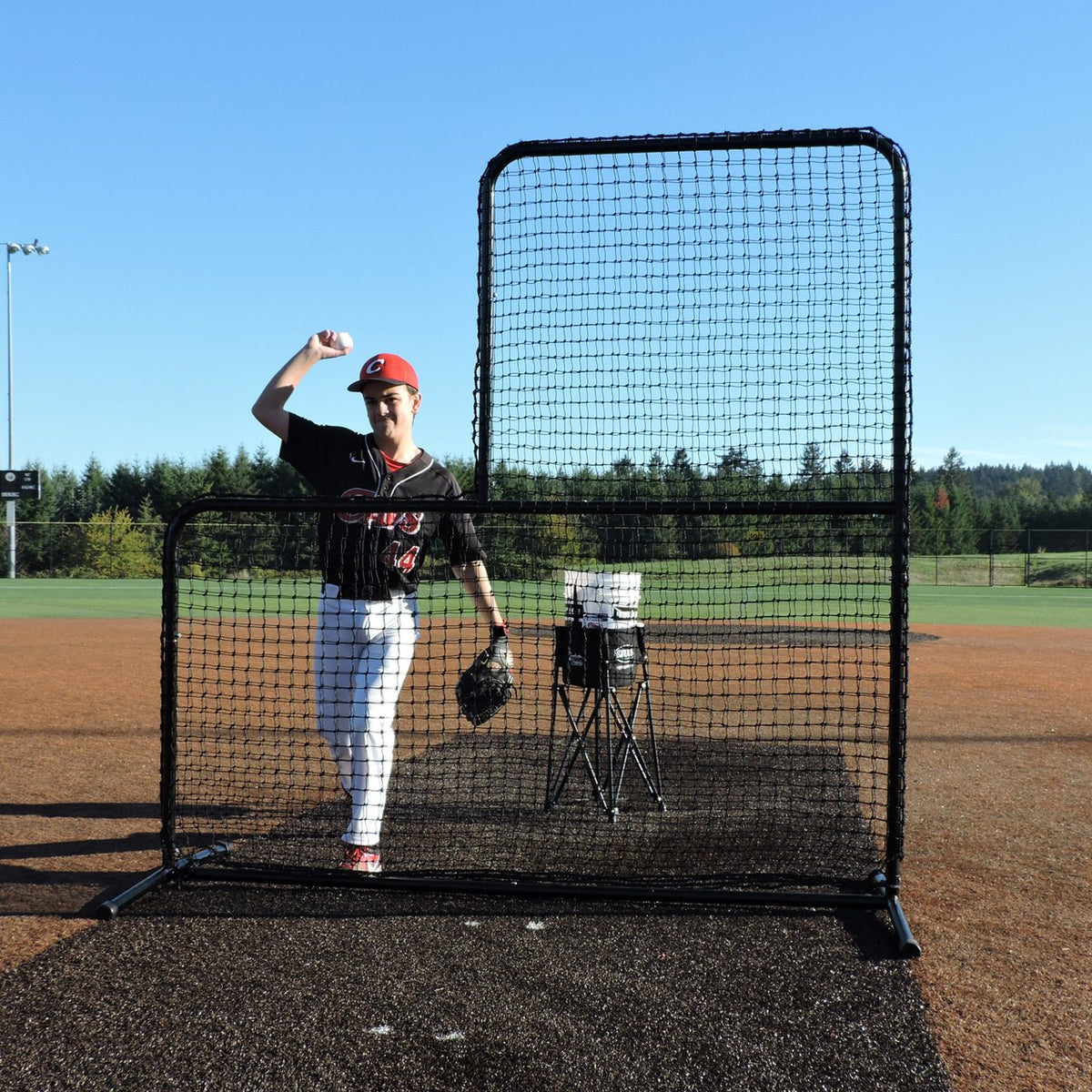 A youth pitcher throwing from behind the JUGS Standard L-Shaped Pitchers Screen on a dirt mound.