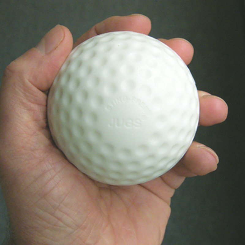 A hand holding a white dimpled practice baseball with JUGS embossed on it, against a dark background.