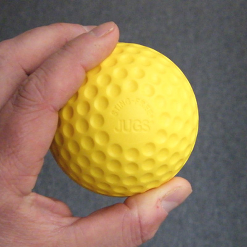 A hand holding a yellow dimpled practice baseball with JUGS embossed on it, against a dark background.