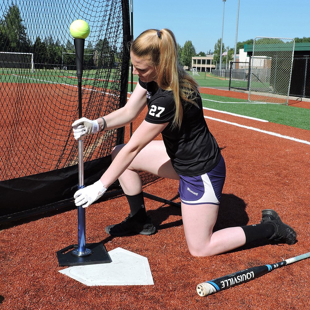 A softball player kneeling to adjust the height of the JUGS T on a home plate base.