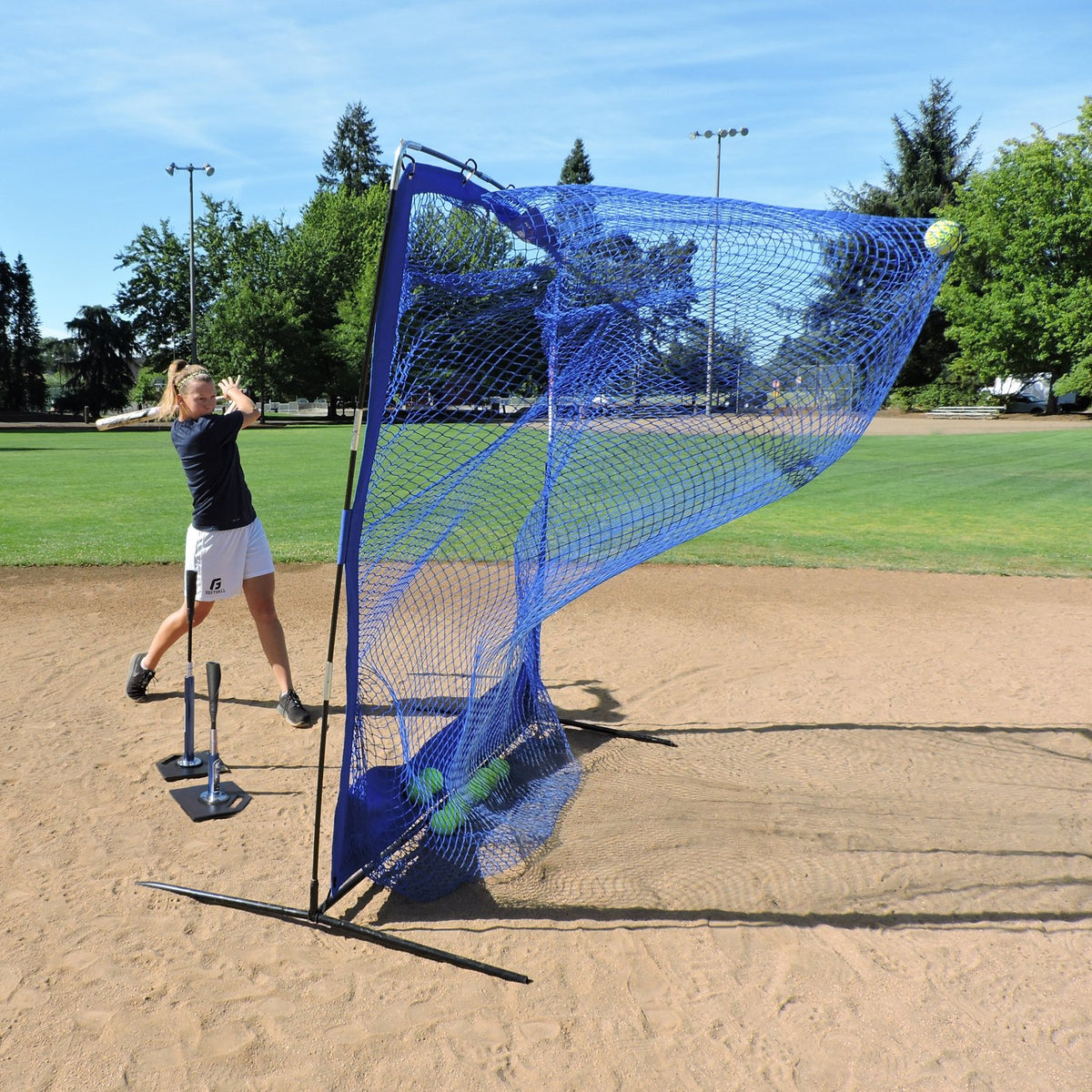 A softball player hitting off the JUGS T into the JUGS Complete Practice Travel Screen on an infield.