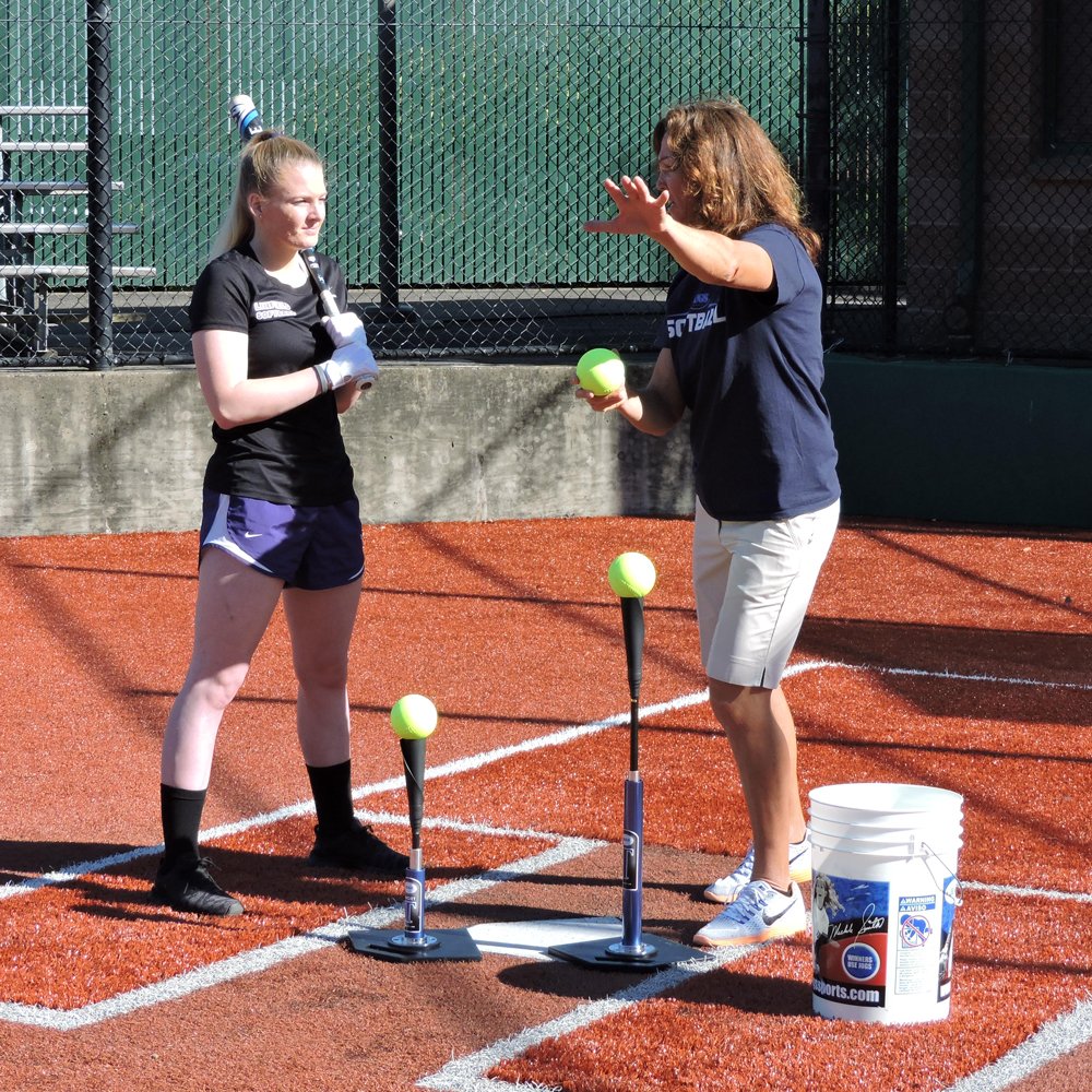 A coach working with a softball hitter at the JUGS T and JUGS Short T on a dirt infield.