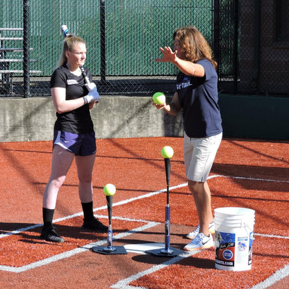 A coach working with a softball hitter at the JUGS T and JUGS Short T on a dirt infield.