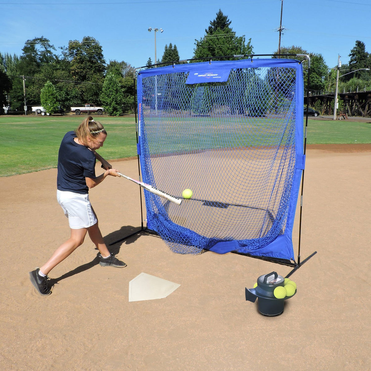 A softball player hitting into the JUGS Complete Practice Travel Screen while the JUGS Toss Machine tosses balls from beside home plate.