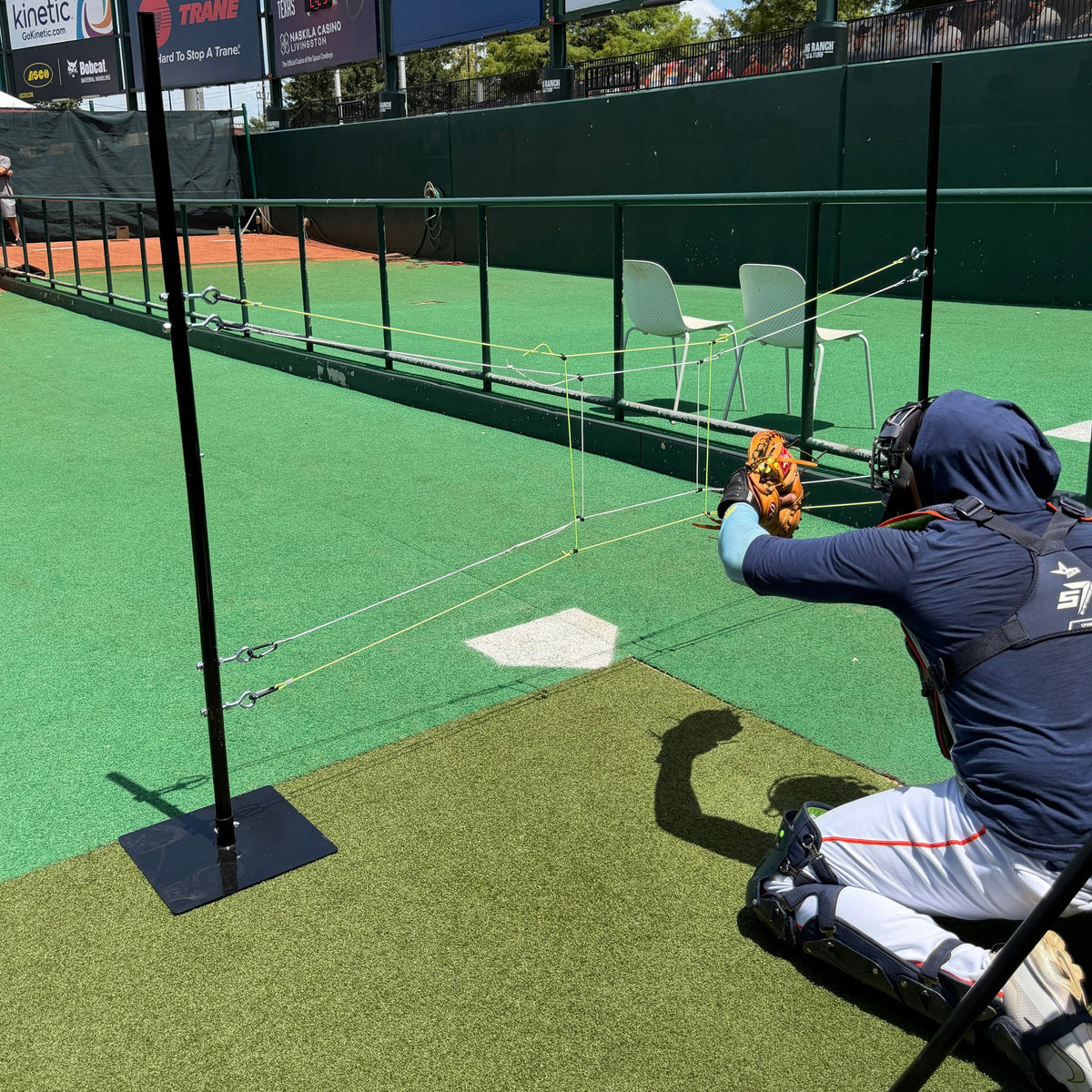 A catcher reaching for a pitch behind a two-pole string target set over home plate in a turf ballpark bullpen