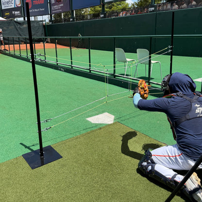 A catcher reaching for a pitch behind a two-pole string target set over home plate in a turf ballpark bullpen