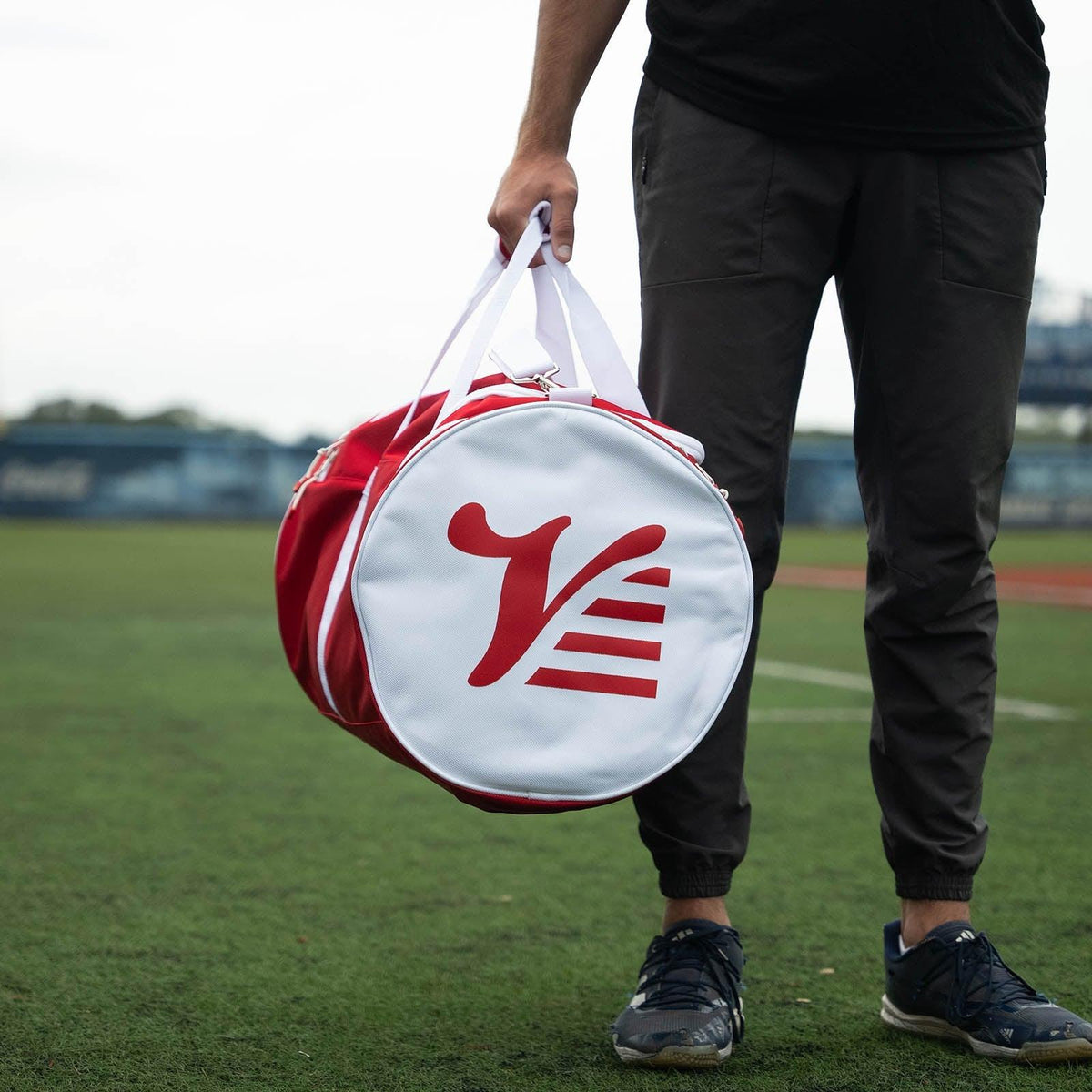 Player holding the red Valle Eagle Players Duffle at a grass baseball field, round logo end cap facing the camera