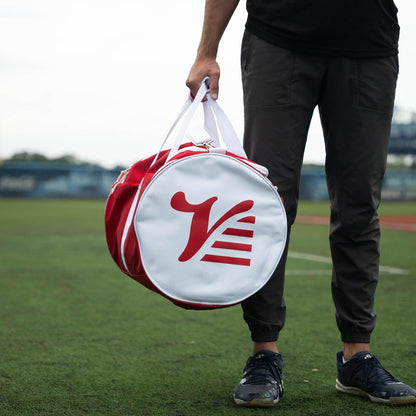 Player holding the red Valle Eagle Players Duffle at a grass baseball field, round logo end cap facing the camera