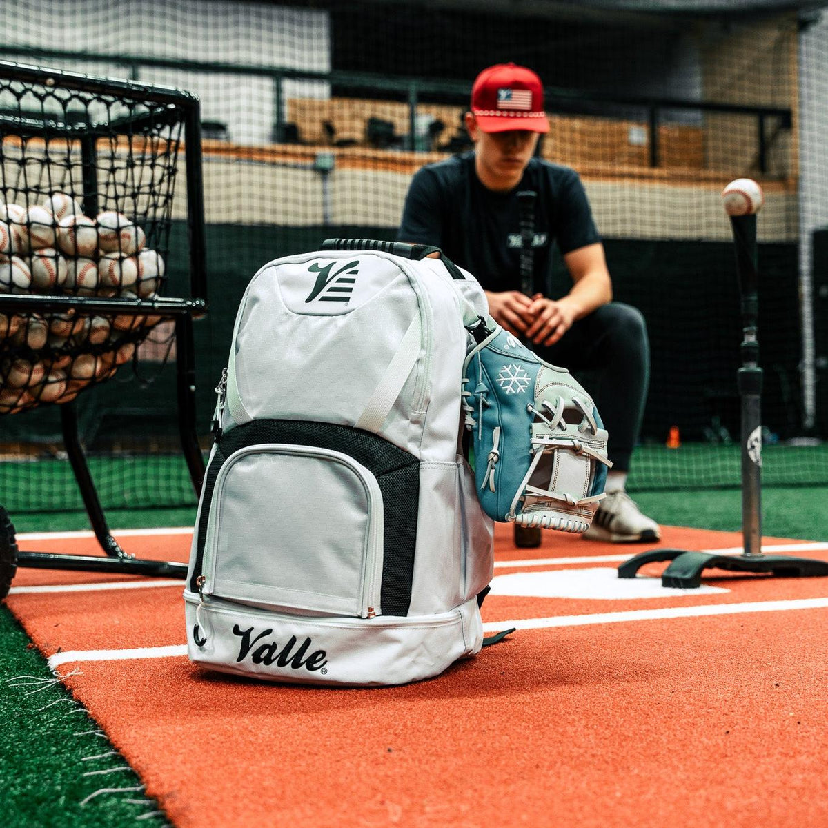Valle Eagle White Baseball Backpack in a batting cage beside a ball caddy, with a training glove resting on it