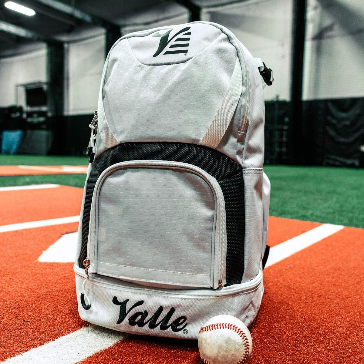 Valle Eagle White Baseball Backpack standing on the home plate stripe of an indoor field, baseball beside it